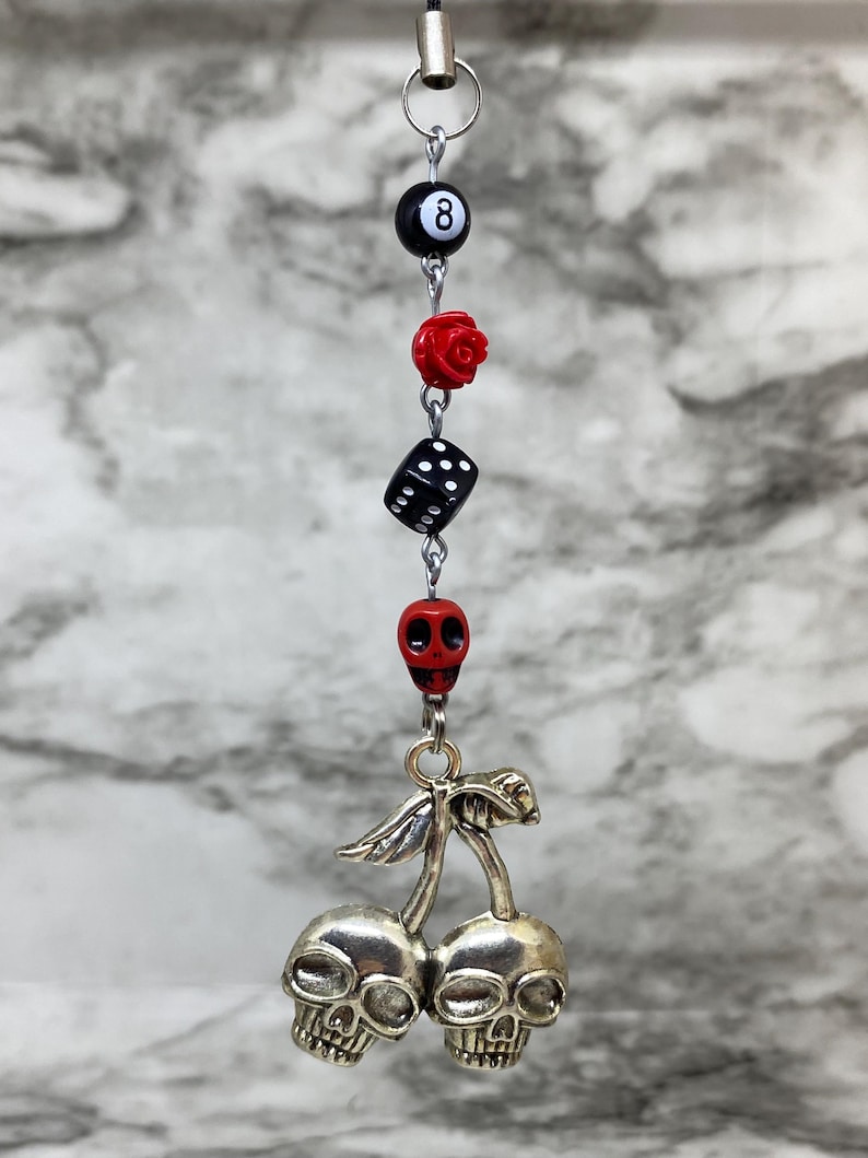 Puede incluir: Un dije colgante con una secuencia vertical de cuentas que incluye una bola ocho negra, una rosa roja, un dado negro y una calavera roja. En la parte inferior, un colgante de metal plateado representa dos calaveras unidas como cerezas.
