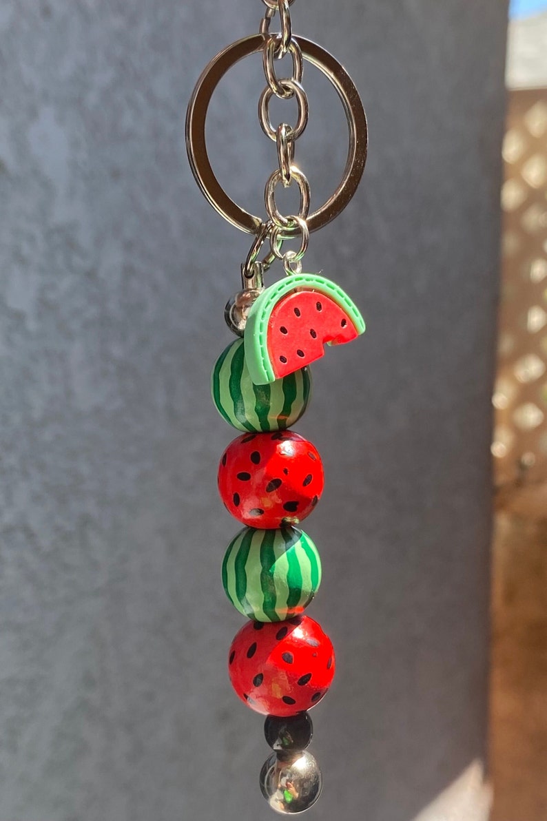 May include: A watermelon keychain with a silver key ring. The keychain features a watermelon slice charm and three round watermelon beads.