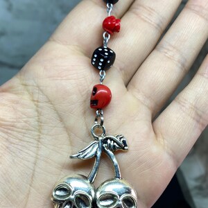 Puede incluir: Un colgante con forma de cerezas de metal plateado donde las cerezas tienen forma de calaveras. La cadena incluye una cuenta negra de bola ocho, una cuenta de calavera roja, un dado negro y otra cuenta de calavera roja unidas por anillas plateadas.