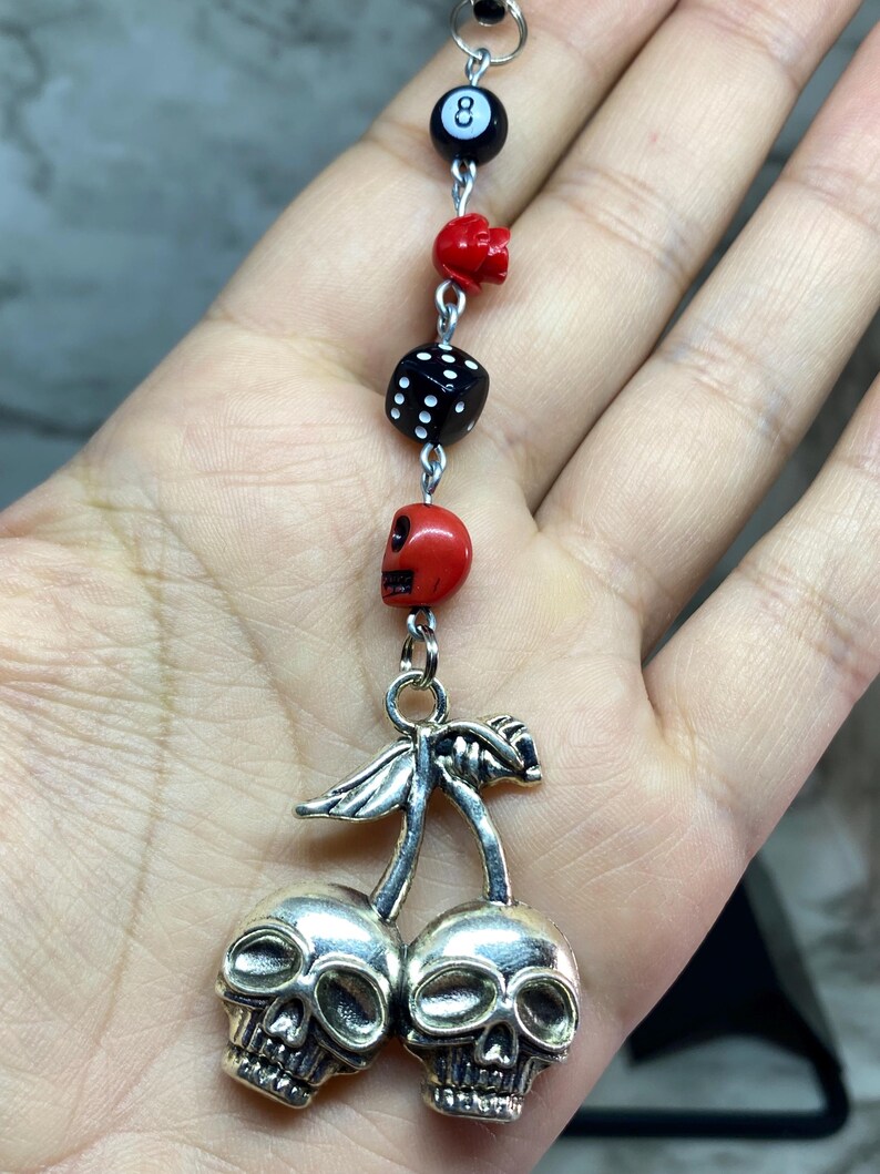 Puede incluir: Un colgante con forma de cerezas de metal plateado donde las cerezas tienen forma de calaveras. La cadena incluye una cuenta negra de bola ocho, una cuenta de calavera roja, un dado negro y otra cuenta de calavera roja unidas por anillas plateadas.