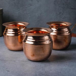 May include: Three copper pots with a polished finish. The pots have a rounded body with horizontal bands and a flared rim. The interior of the pots reflects light, creating a warm glow. The pots are arranged on a grey surface.