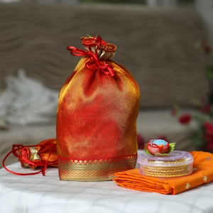 Puede incluir: Una bolsa roja y dorada con cordón y ribete dorado, un pequeño recipiente transparente con brazaletes dorados y una tela naranja doblada. Una pequeña flor roja y azul se encuentra encima del recipiente.