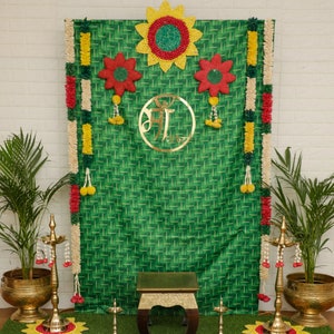 Pode incluir: Um fundo verde com um padrão tecido decorado com grinaldas florais amarelas, vermelhas e brancas. Um círculo dourado com a palavra "Aarti" em escrita hindi está centralizado no fundo. Um pequeno banquinho dourado fica na frente do fundo.