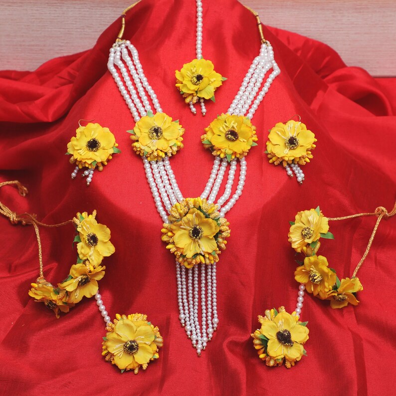 May include: A yellow flower jewelry set with a pearl necklace, earrings, bangles, and a maang tikka. The jewelry is on a red fabric background.