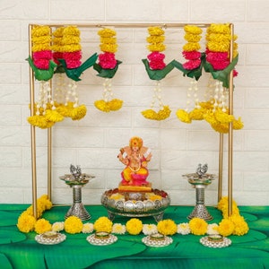 May include: A decorative gold metal frame adorned with yellow and pink floral garlands, green leaf accents, and strings of white beads. A colorful statue of Ganesha sits on a silver pedestal, flanked by silver lamps and candles.