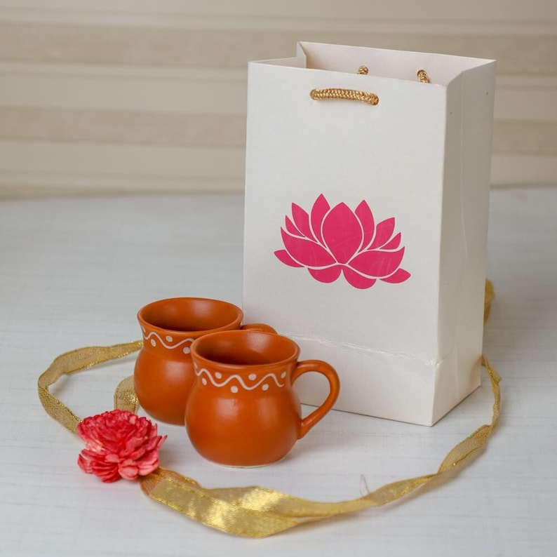 Puede incluir: Dos tazas de terracota con dise&ntilde;os de ondas blancas junto a una bolsa de regalo blanca con una flor de loto rosa. Tambi&eacute;n hay una cinta dorada y una flor roja. Las tazas son peque&ntilde;as, perfectas para t&eacute; o caf&eacute;.
