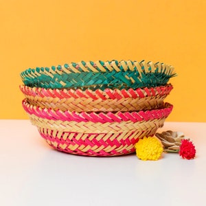 May include: A stack of three woven baskets in shades of green, red, and pink. The baskets are round with a braided design. A yellow pom-pom and a small decorative flower are next to the baskets against a yellow background.