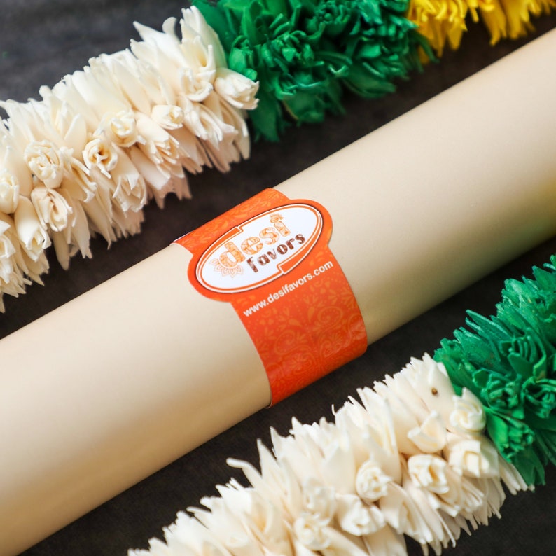 May include: A white, cream, and green garland of artificial flowers with a brown paper tube wrapped in orange paper with the text "desi favors" and the website "www.desifavors.com".
