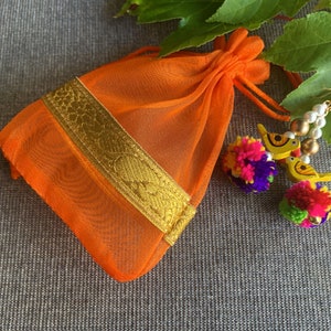 May include: An orange mesh drawstring bag with a gold trim. The bag is sitting on a gray textured surface with colorful pom-poms and wooden bird ornaments.