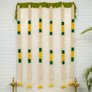 May include: A set of seven white, yellow, and green flower garlands hanging on a white wall. The garlands are made of artificial flowers and are used for decoration.