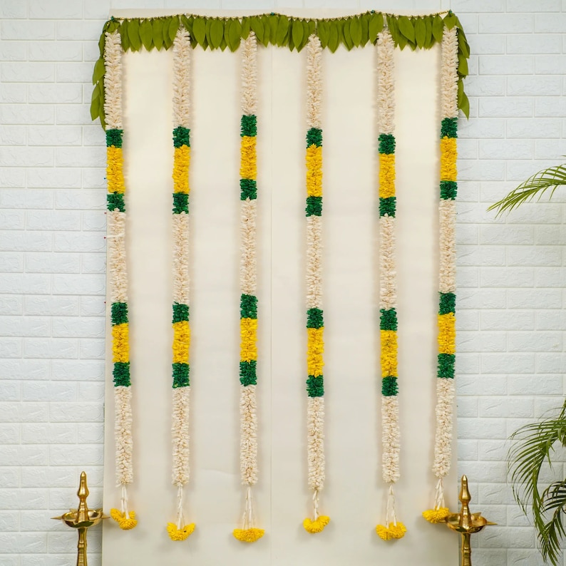 May include: A set of seven white, yellow, and green flower garlands hanging on a white wall. The garlands are made of artificial flowers and are used for decoration.