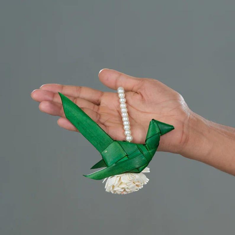 May include: A green bird made of woven palm leaves with a white flower attached to the bottom. A string of white pearls hangs from the bird's neck.