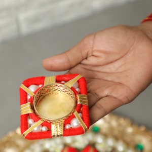May include: A red and gold square-shaped candle holder with white pearl accents. The holder has a round, gold metal insert for a tea light candle.