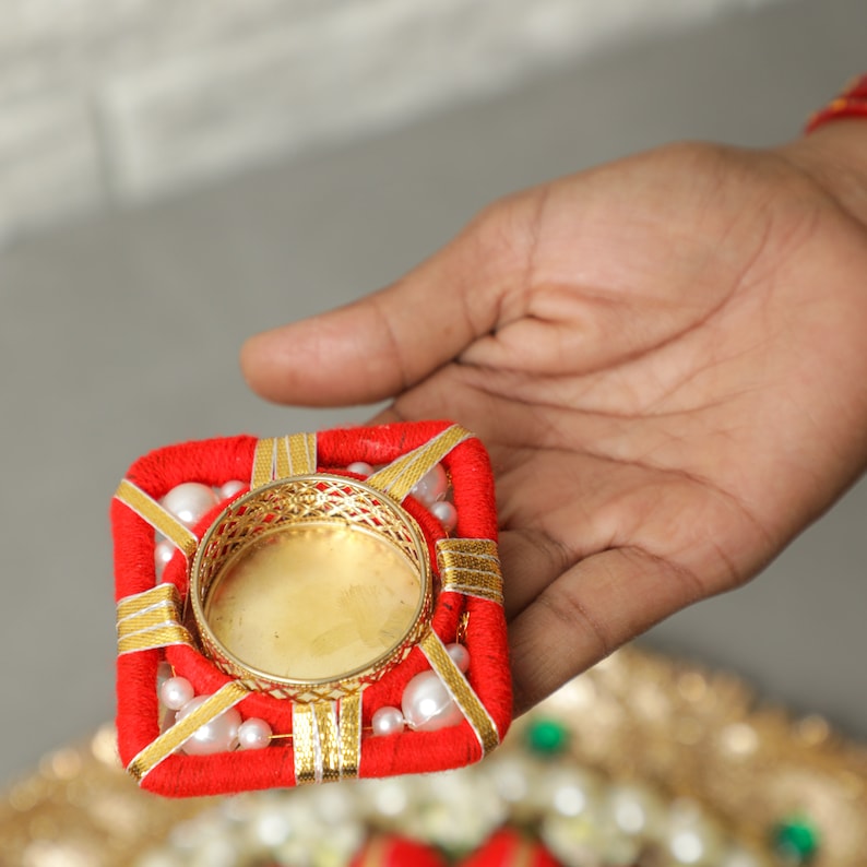 May include: A red and gold square-shaped candle holder with white pearl accents. The holder has a round, gold metal insert for a tea light candle.