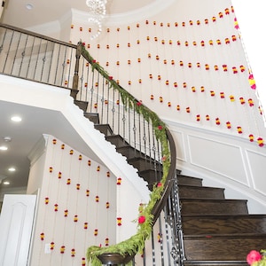 May include: A staircase decorated with strings of red, yellow, and white flower garlands. Green garland adorns the banister. The stairs are made of dark wood, and the walls are painted white. A chandelier hangs from the ceiling.
