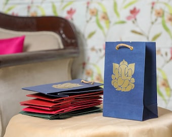 Può includere: Un mucchio di sacchetti regalo blu, rossi e verdi con un design Ganesha dorato. I sacchetti sono su un tavolo marrone chiaro. Un sacchetto regalo blu con un design Ganesha dorato è in primo piano.