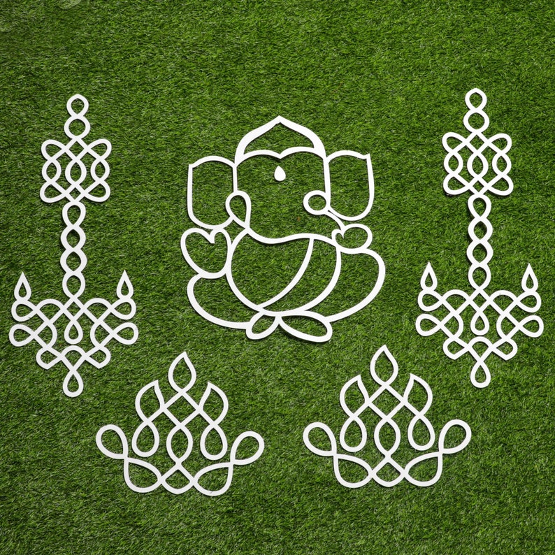 May include: White paper cutouts of traditional Indian designs, including a depiction of the Hindu deity Ganesha, on a green grass background.