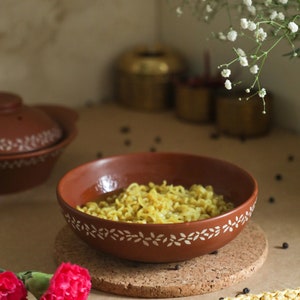 Puede incluir: Un bol de terracota con un diseño floral blanco lleno de fideos amarillos y un caldo claro. El bol está sobre un posavasos de corcho.
