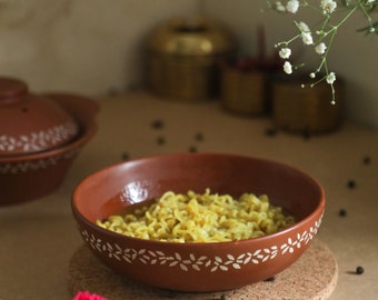 May include: A terracotta bowl with a white floral design filled with yellow noodles and a clear broth. The bowl is sitting on a cork coaster.