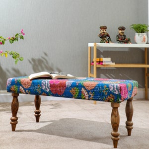 Może przedstawiać: Prostokątna ławka z kolorową poduszką w stylu patchwork w odcieniach niebieskiego, różowego i pomarańczowego. Ławka ma cztery toczone drewniane nogi. Otwarta książka i okulary spoczywają na poduszce. W tle widoczne są figurki dekoracyjne i roślina.