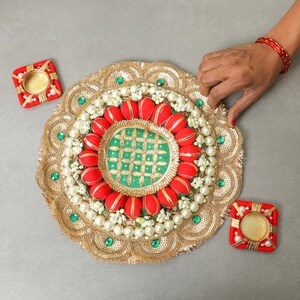 May include: A gold circular decorative plate with red and green fabric flowers, white pearls, and green gems. Two small red square candle holders are on either side of the plate.