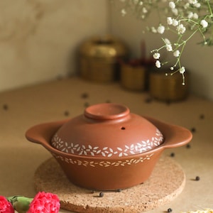 May include: A terracotta cooking pot with a lid, featuring a white leaf design. The pot is sitting on a cork trivet.