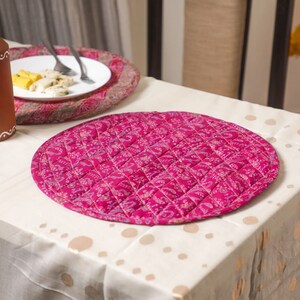 May include: A round pink quilted placemat with a floral pattern. The placemat is on a white tablecloth with gold polka dots.