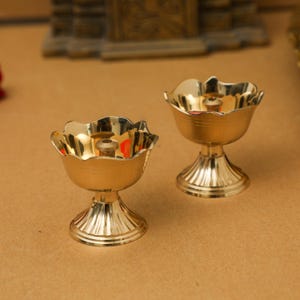 Puede incluir: Dos lámparas de aceite de metal dorado con base acanalada y borde festoneado. Las lámparas están sobre una superficie marrón.