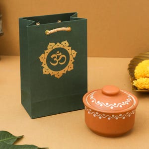 May include: A dark green gift bag with a gold Om symbol and rope handles stands next to a small, round, terracotta-colored pot with a lid. The pot has a white dotted design around the rim.
