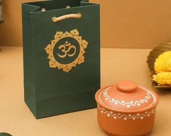 May include: A dark green gift bag with a gold Om symbol and rope handles stands next to a small, round, terracotta-colored pot with a lid. The pot has a white dotted design around the rim.