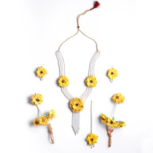 May include: A yellow flower jewelry set with a pearl necklace, earrings, a maang tikka, and two bangles. The necklace has three strands of pearls with yellow flower accents. The earrings are small and feature a single yellow flower. The maang tikka is a long, thin chain with a yellow flower at the end. The bangles are made of pearls and have yellow flower accents.