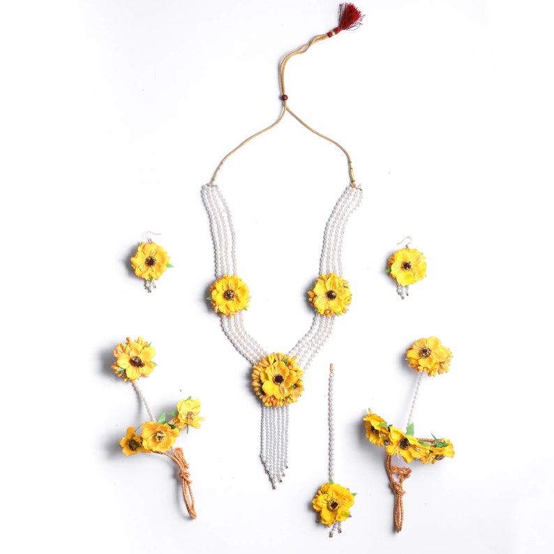 May include: A yellow flower jewelry set with a pearl necklace, earrings, a maang tikka, and two bangles. The necklace has three strands of pearls with yellow flower accents. The earrings are small and feature a single yellow flower. The maang tikka is a long, thin chain with a yellow flower at the end. The bangles are made of pearls and have yellow flower accents.