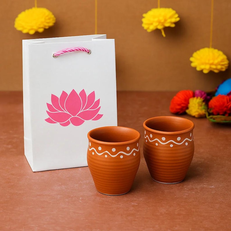 Puede incluir: Una bolsa de regalo blanca con un dise&ntilde;o de flor de loto rosa y dos tazas de color terracota con patrones de ondas punteadas blancas. Pompones amarillos en el fondo sugieren un tema festivo.