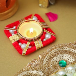 May include: A red and gold decorative candle holder with white beads and a lit tea light candle in the center.