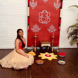 May include: A young woman in a beige and gold dress sits in front of a red and white decorated backdrop. The backdrop features a traditional Indian design with garlands of flowers and leaves. The woman is smiling and looking at the camera. There are several small plates of food on the floor in front of her.