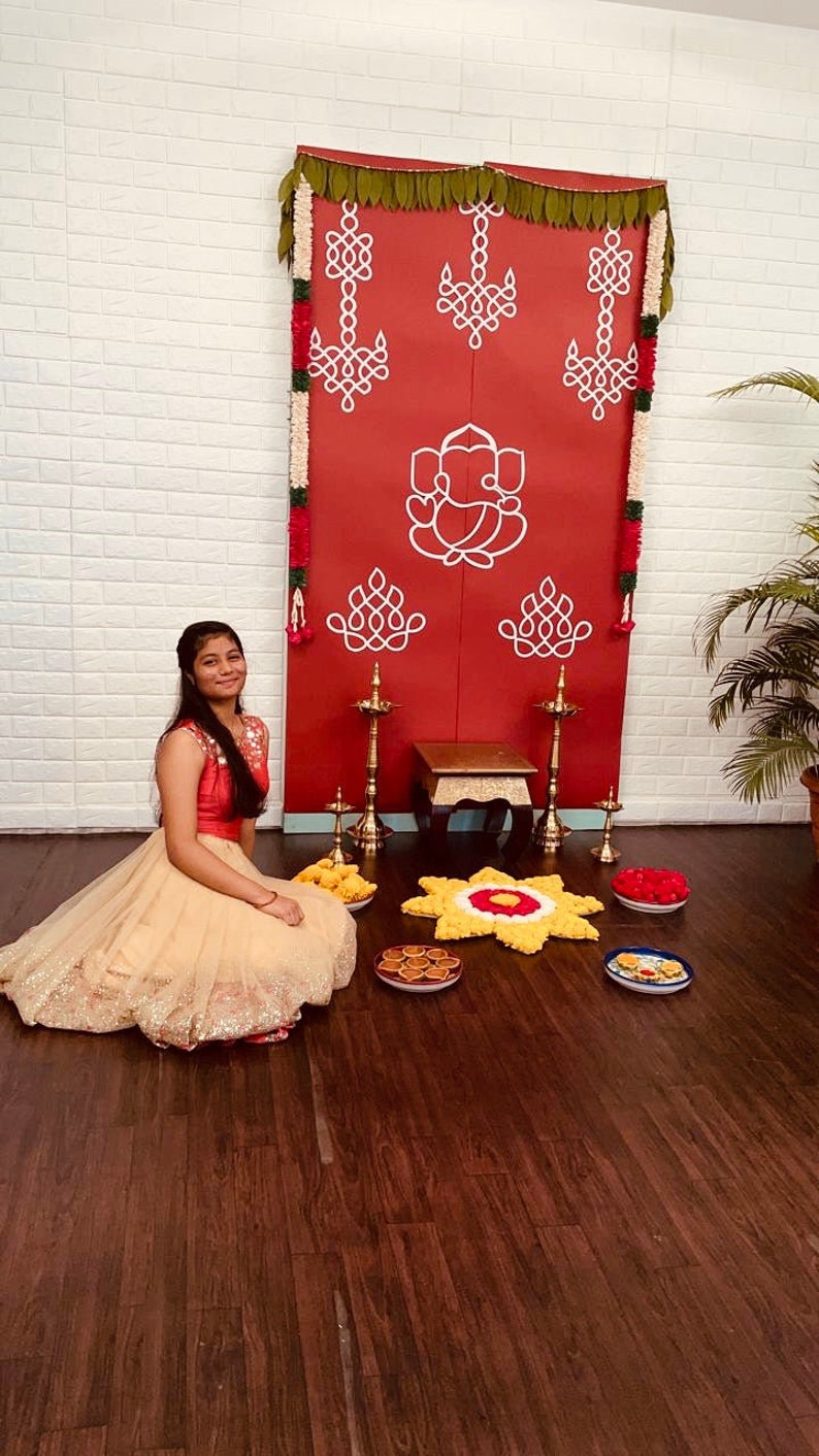 May include: A young woman in a beige and gold dress sits in front of a red and white decorated backdrop. The backdrop features a traditional Indian design with garlands of flowers and leaves. The woman is smiling and looking at the camera. There are several small plates of food on the floor in front of her.