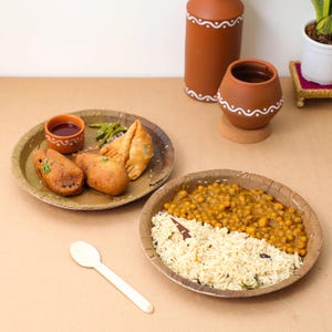 May include: A table setting featuring Indian cuisine. Two plates hold various dishes, including samosas, fried snacks, and a chickpea and rice dish. A wooden spoon and small terracotta pots with white painted designs complete the scene.
