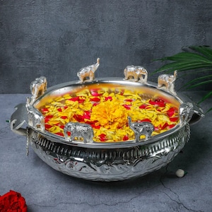 Può includere: Una ciotola d'argento con intricati disegni floreali, riempita d'acqua, petali di fiori gialli e rossi e un singolo fiore giallo. Otto figurine di elefanti d'argento sono posizionate attorno al bordo della ciotola.