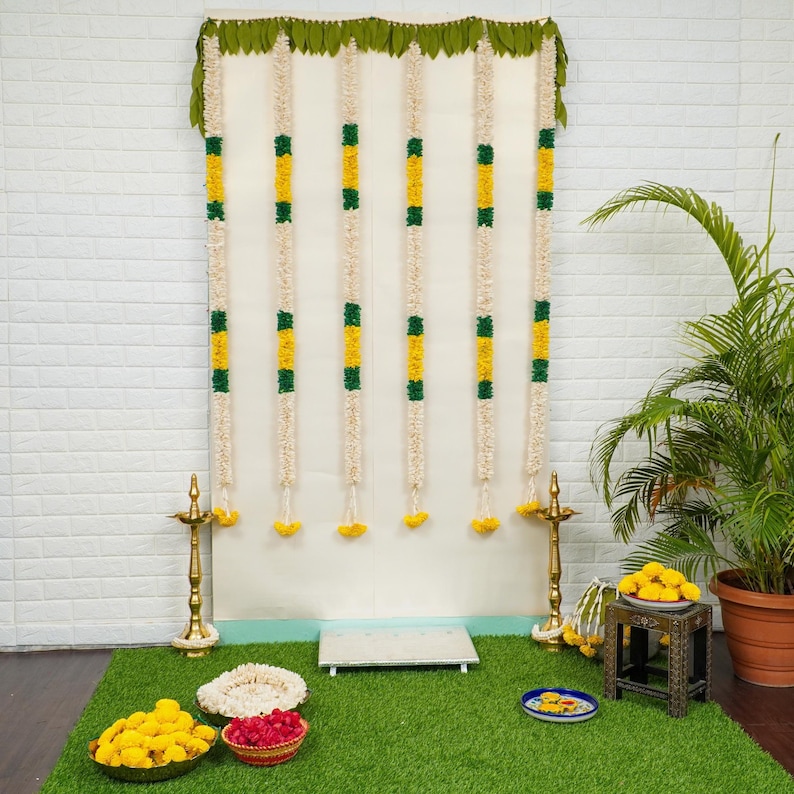 May include: A white backdrop decorated with green and yellow flower garlands, two gold candle holders, and a small platform. The backdrop is set up on a green artificial grass surface with yellow flowers, red beads, and a small plate.