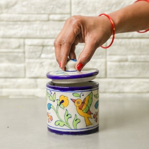 May include: A hand-painted ceramic jar with a lid, decorated with a bird, flowers, and leaves in yellow, green, and blue. The jar has a white base with blue trim. The lid is being lifted, revealing the inside of the jar.