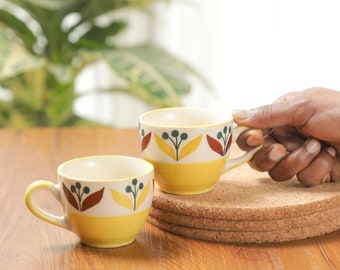 Op de afbeelding: Twee gele keramische mokken met bruine en groene bloemenmotieven. De mokken staan op een houten tafel met een kurken onderzetter.