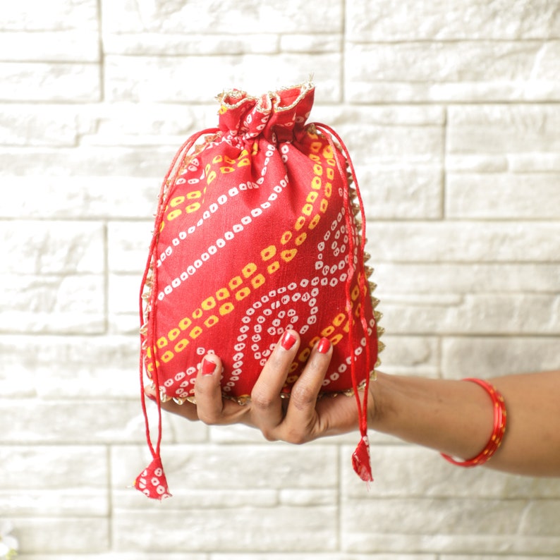 Pu&ograve; includere: Una borsa a cordoncino rossa con un motivo bianco e giallo. La borsa &egrave; decorata con perline dorate e ha un fiocco.