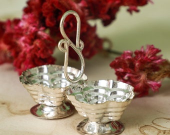 May include: A silver-coloured, two-part serving dish with a handle. The dish has a scalloped edge and sits on a small pedestal base. The handle is a decorative, looped design. Red flowers are in the background.