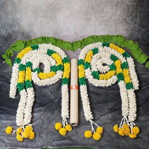 May include: Two white flower garlands with green and yellow accents, each with a tassel of yellow flowers at the end. The garlands are arranged in a V-shape with a brown paper roll in the center.