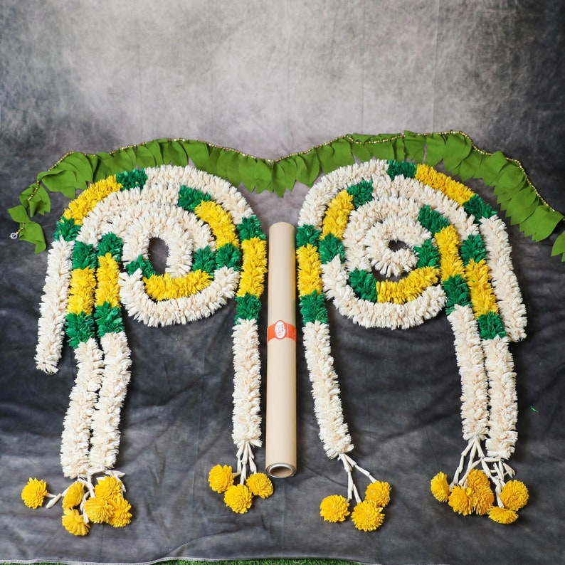 May include: Two white flower garlands with green and yellow accents, each with a tassel of yellow flowers at the end. The garlands are arranged in a V-shape with a brown paper roll in the center.
