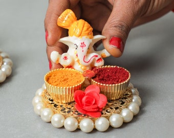 May include: A white Ganesha figurine with a yellow headdress sits on a gold tray with two small bowls filled with yellow and red powder. The tray is surrounded by a pearl-like border.