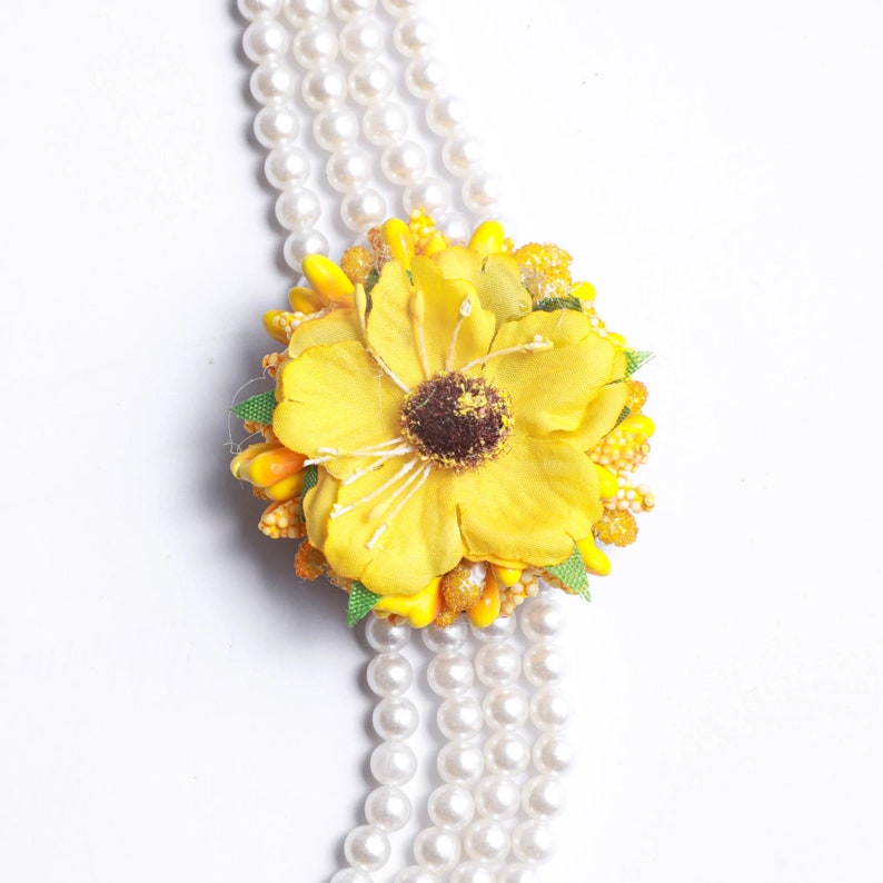 May include: A yellow flower with a brown center is attached to a strand of white pearls. The flower is surrounded by small yellow and orange beads.