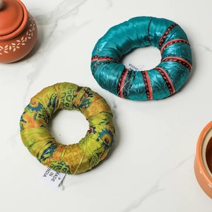 May include: Two decorative ring-shaped objects. One is teal with red and black accents, the other is yellow and green with blue and orange patterns. Both are made of fabric. The rings are on a white surface, near a small orange pot.