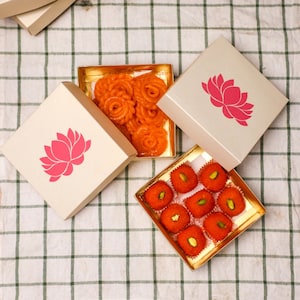 May include: Three boxes with different Indian sweets. The boxes are decorated with a pink lotus flower design. The sweets are arranged in a square shape. The sweets are orange and red.