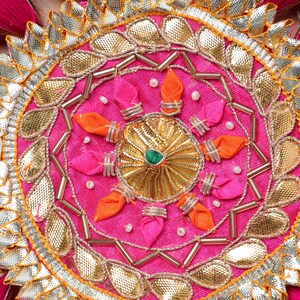 May include: A pink fabric circle with a gold and silver design. The center of the circle has a gold and green flower design. The circle is decorated with small pink and orange fabric flowers.